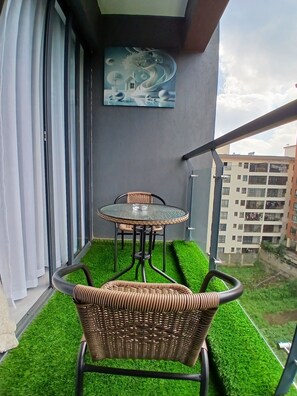Ein Balkon mit Kunstgrashimmel, einem Korbstuhl, einem Glastisch und einem Gemälde.