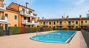 Une piscine entourée d’immeubles jaunes à plusieurs étages, dotés de balcons, sous un ciel dégagé, et bordée d’une haie.
