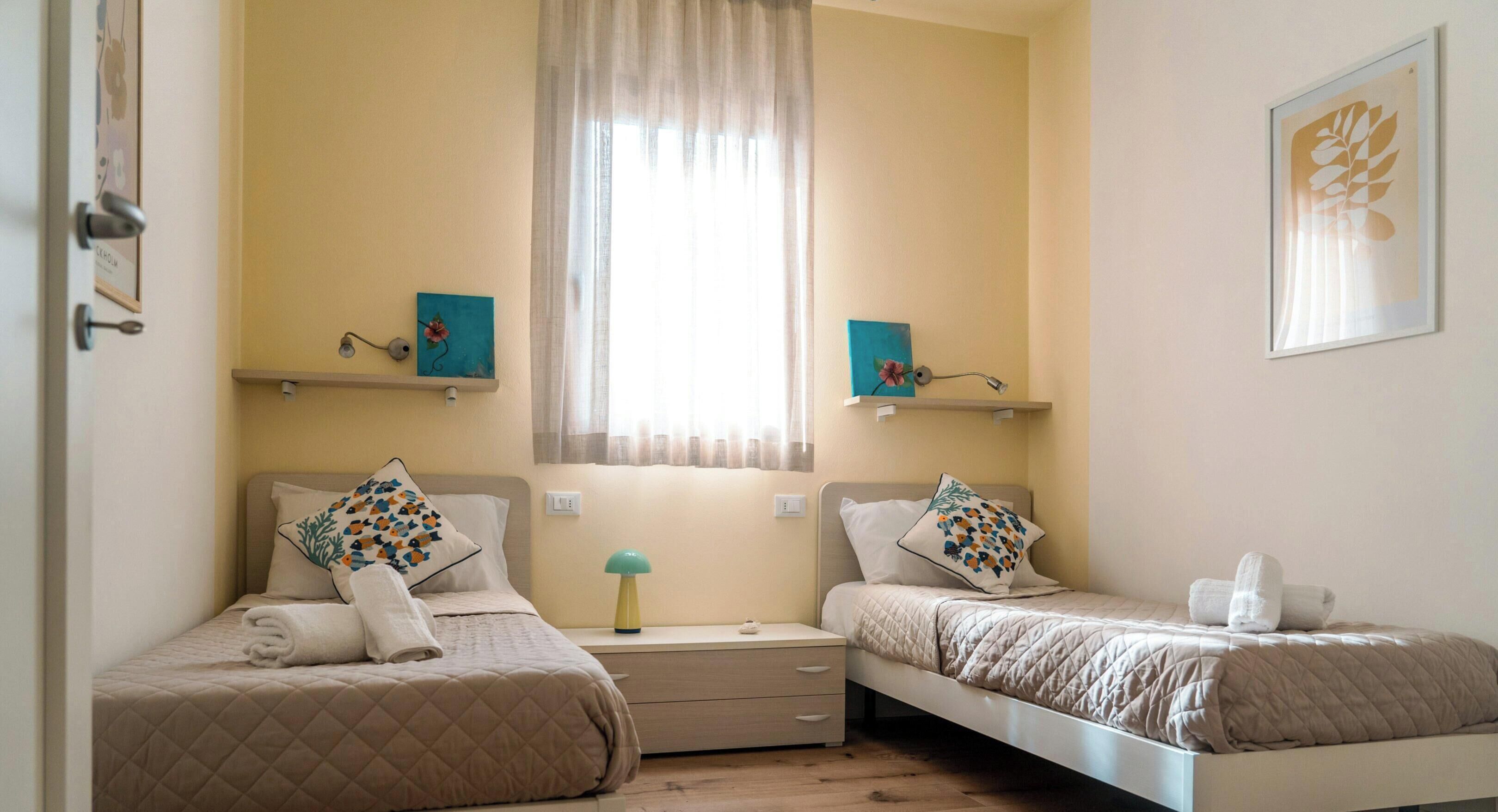 A bedroom with two beds, a window with curtains, and artwork on the walls.