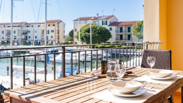 Uma mesa de madeira com pratos, copos e um jarro, com vista para uma marina e edifícios ao fundo.