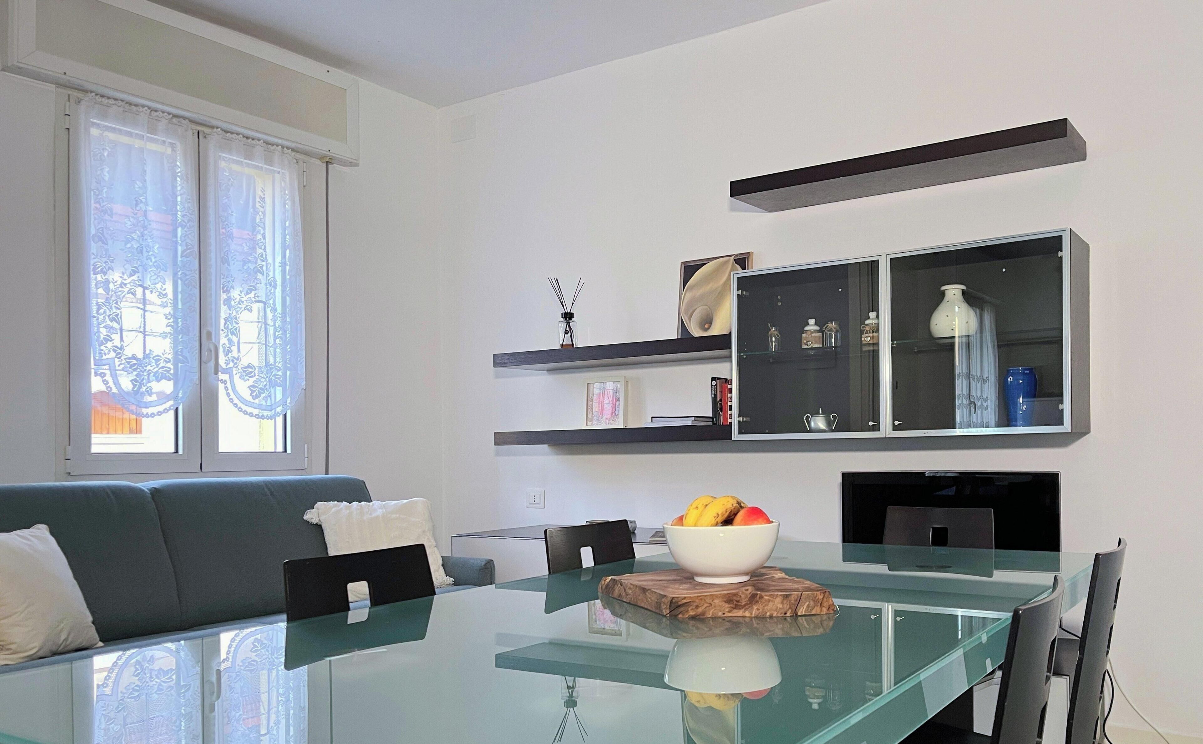 A modern living room with a glass dining table, a sofa, a TV, and a kitchen area with shelves.