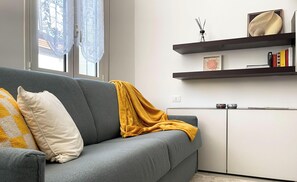 Un sofá gris con una manta amarilla, una almohada blanca y una almohada amarilla con estampado de cuadros. Hay dos estantes con libros y objetos decorativos.