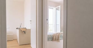 A narrow hallway leading to a bedroom with a window, a bedside table with a lamp, and a bed with a patterned bedspread. - SkyLux, 2-bedroom – 4th floor with view (Lido di Iesolo)
