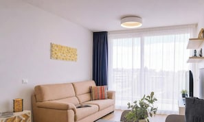 A living room with a sofa, a potted plant, a flat-screen TV, and a window with curtains. - SkyLux, 2-bedroom – 4th floor with view (Lido di Iesolo)