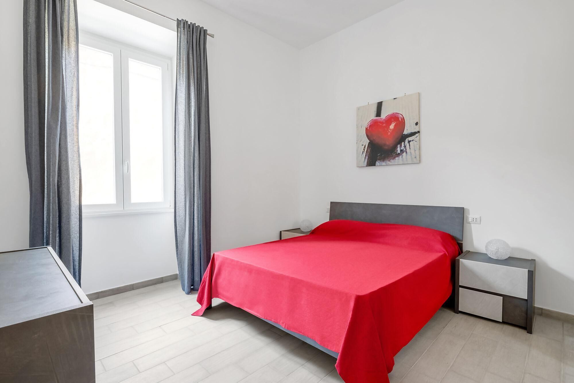 A bedroom with a bed, a nightstand, a bedside table, a window with curtains, and a painting on the wall.