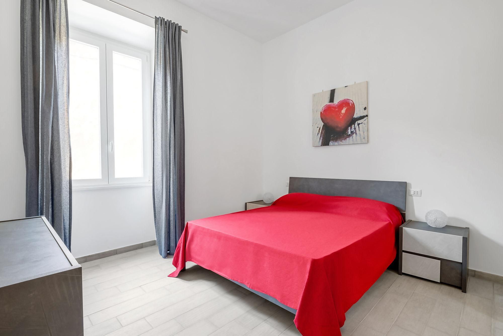 A bedroom with a bed, a nightstand, a bedside table, a window with curtains, and a painting on the wall.