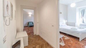 Quarto de hotel com uma cama, uma mesa pequena e uma cadeira. Há um corredor com uma porta que dá para outro quarto.