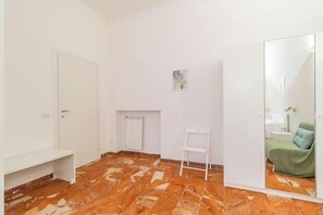 Une pièce avec un sol carrelé, une porte blanche, un radiateur blanc, une étagère blanche et un miroir qui reflète un lit recouvert d’une couverture verte.
