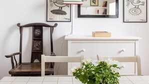 Una habitación con piso de baldosas blancas, una silla de madera, un aparador blanco, una planta en maceta verde y dos cuadros en la pared.