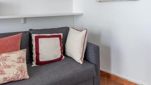 Un sofá gris con cojines estampados, un florero rojo y un retrato en un marco sobre una pared blanca.