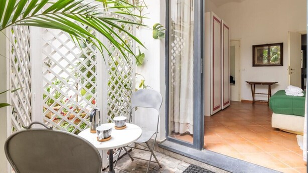 Una habitación con una silla blanca, una mesita redonda pequeña, una cama con cubierta verde y una puerta que da a otra habitación.