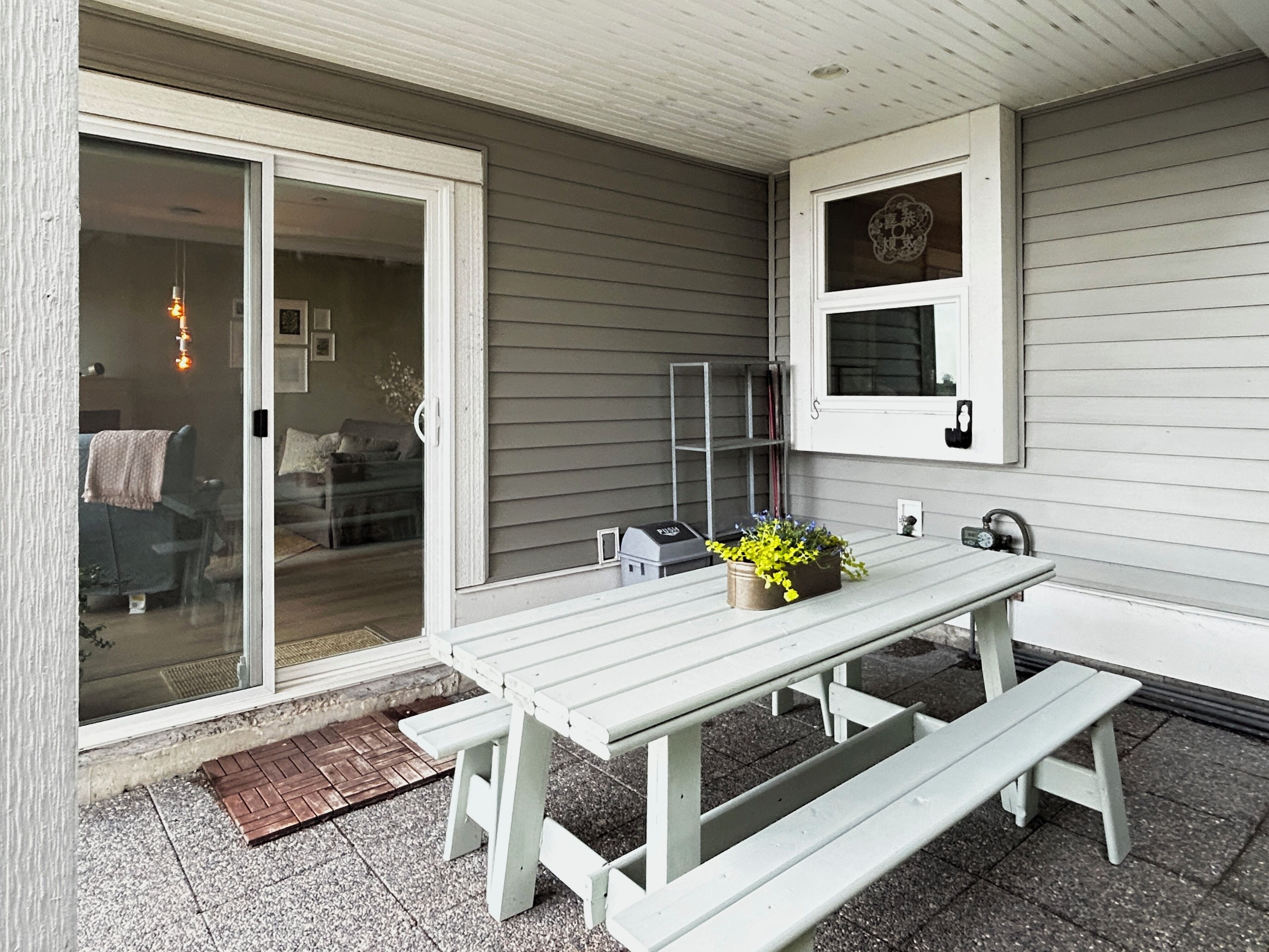 Une terrasse avec une table et des bancs blancs, une porte-fenêtre coulissante, une petite plante en pot et une étagère murale agrémentée d’un objet décoratif.