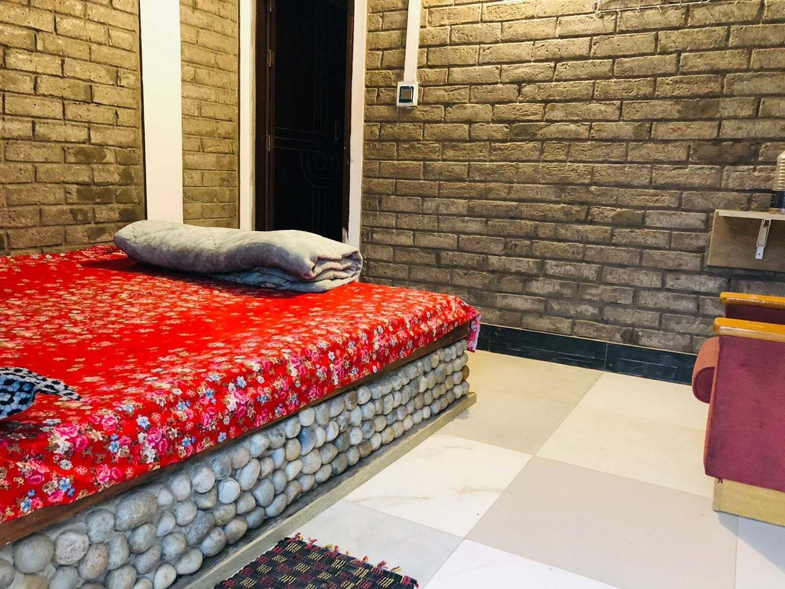 A room with a bed against a brick wall, a window, and a small shelf with items.