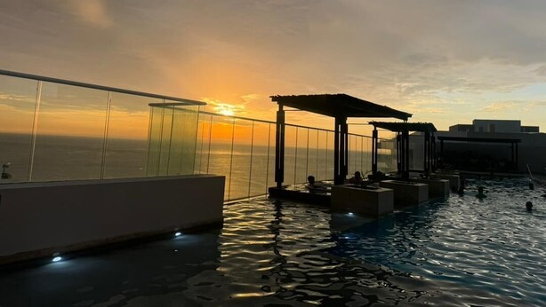 A rooftop infinity pool with a view of the sunset over the ocean, surrounded by glass railings and lounge areas. - Pleasant 1-bedroom apartment with AC in vibrant Rodadero (Rodadero)