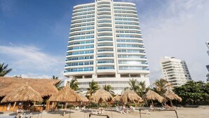 A tall, modern hotel with a curved facade, multiple balconies, and a beachside location with thatched umbrellas and lounge chairs. - Pleasant 1-bedroom apartment with AC in vibrant Rodadero (Rodadero)