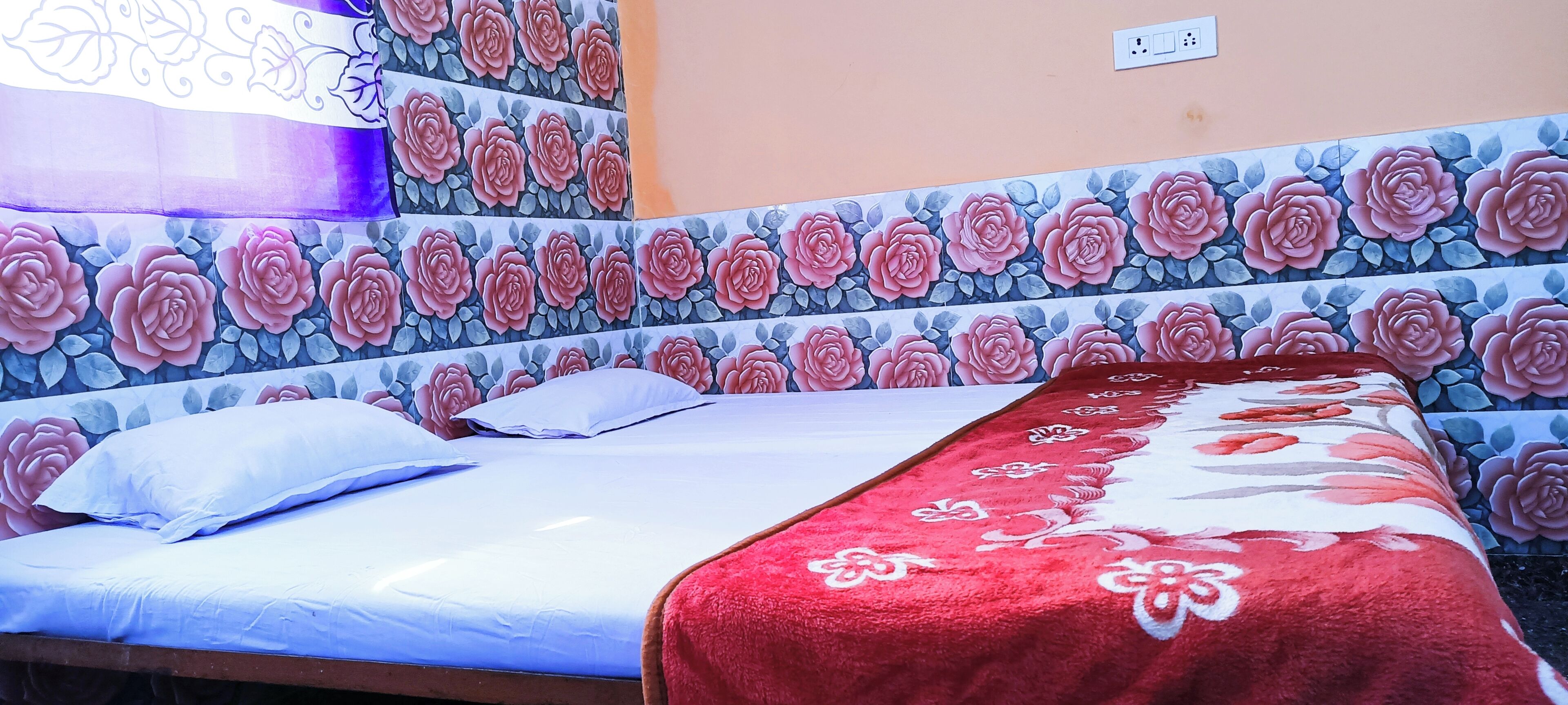 A single bed with a white sheet and a red floral blanket, a white pillow, and a wall with a floral pattern.