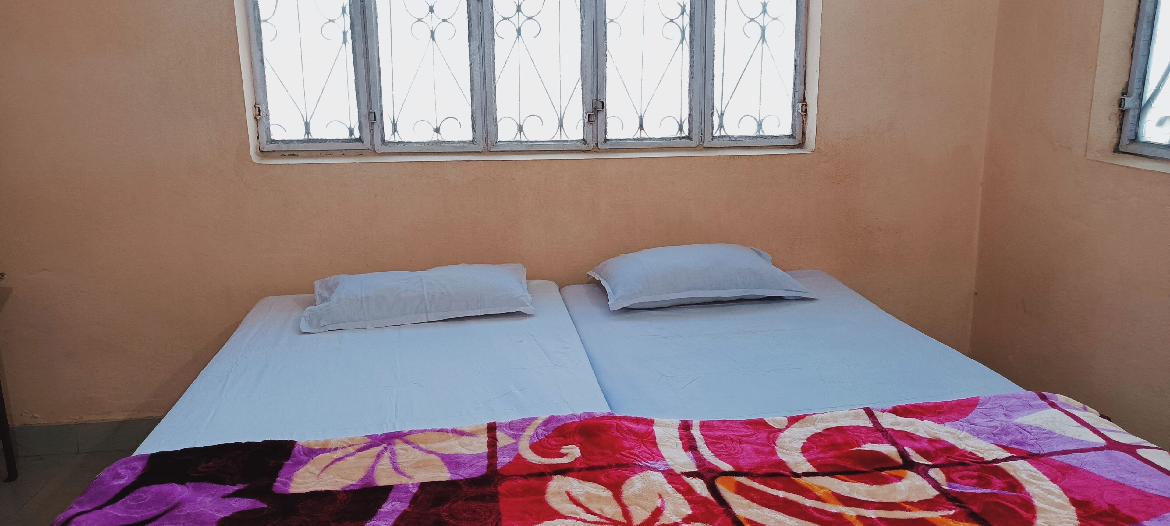 A single bed with two pillows and a colorful blanket, a window with metal bars, and a plain wall.