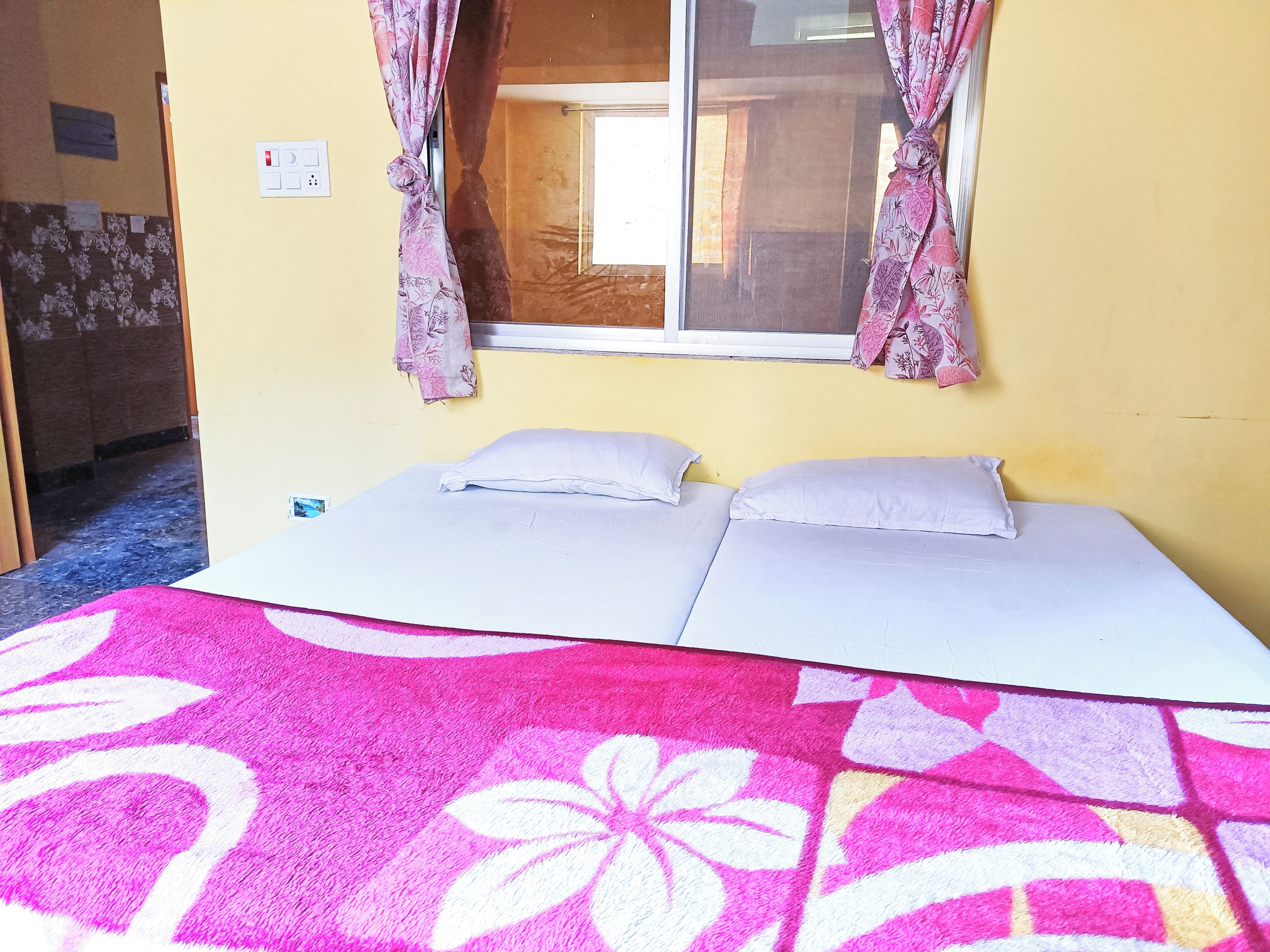 A room with two beds, a window with curtains, a wall-mounted light switch, and a door leading to another room.