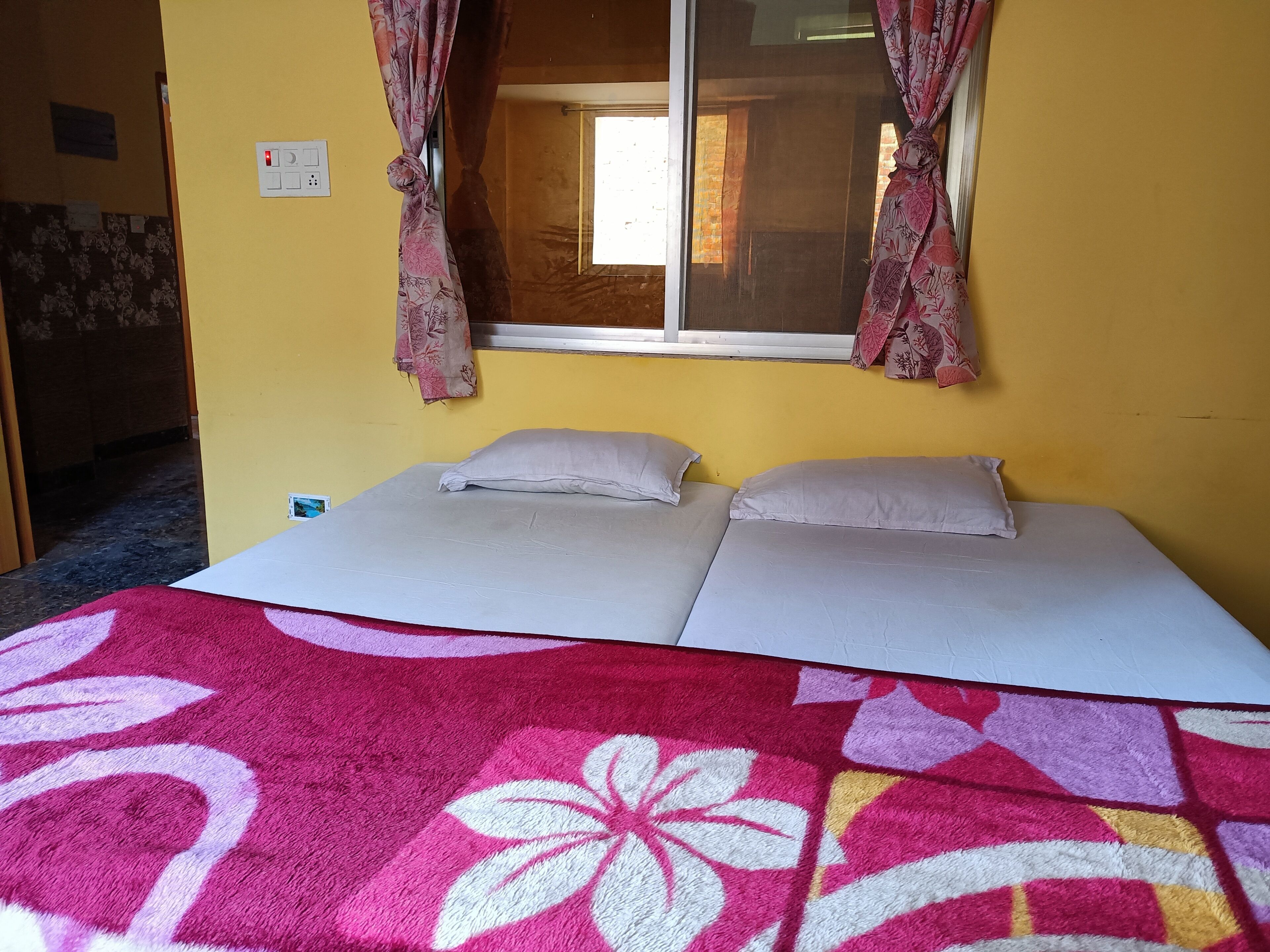 A room with a bed, two pillows, a window with curtains, and a wall socket.
