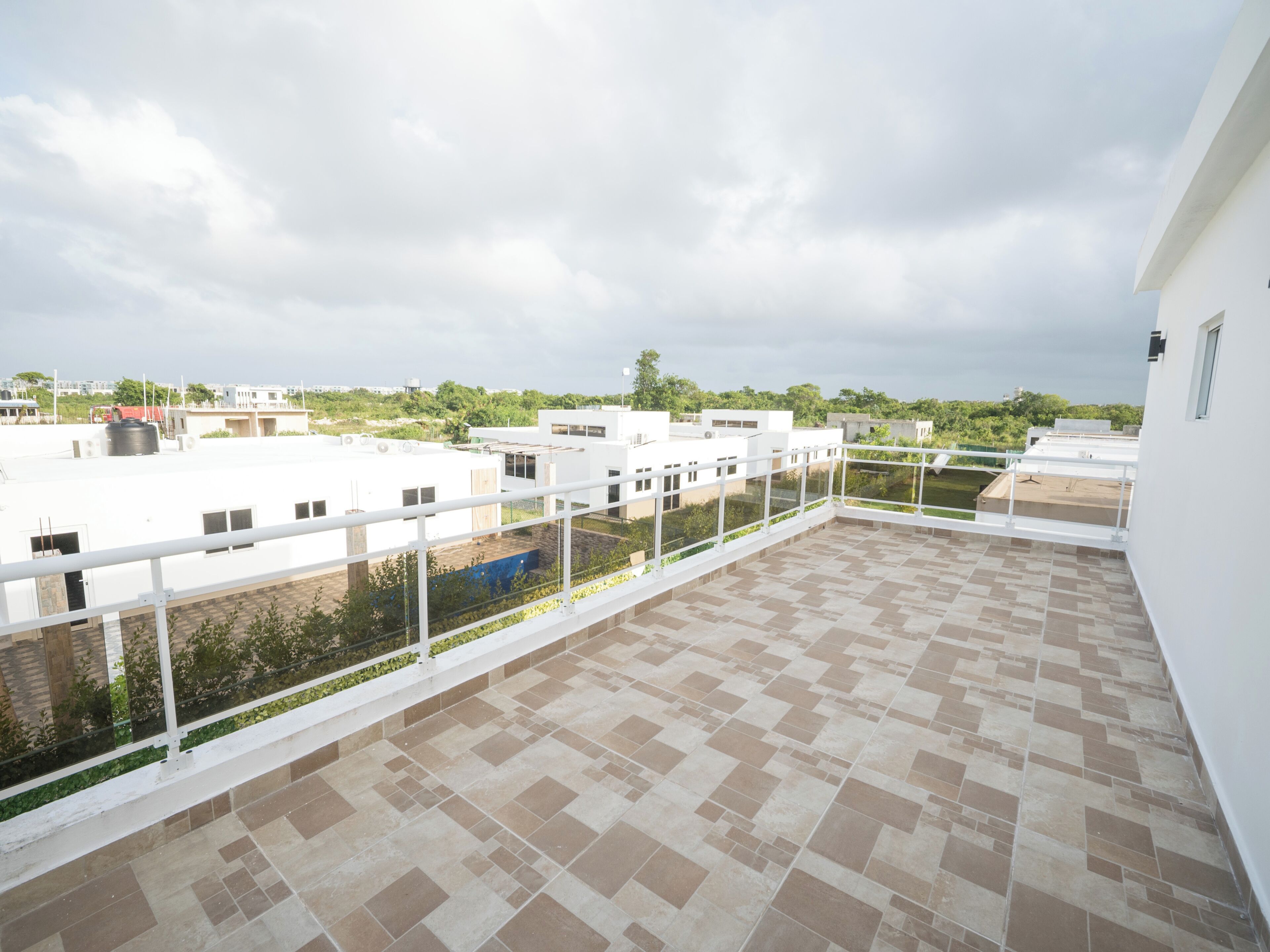 A rooftop terrace with a tiled floor, white railings, and a view of surrounding buildings and greenery.