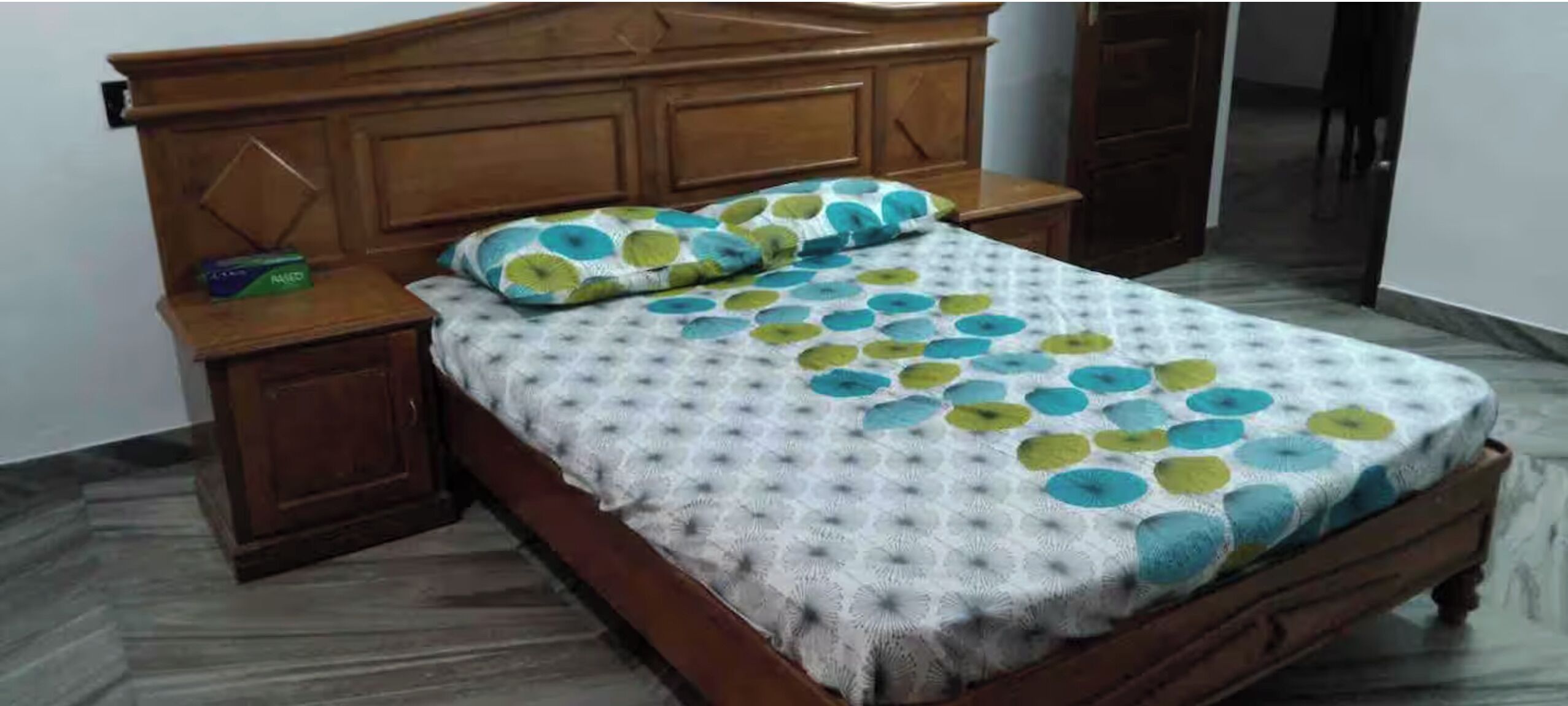 Een houten bed met een bedsprei en kussens met een patroon, een nachtkastje met een doosje, en een houten garderobe.
