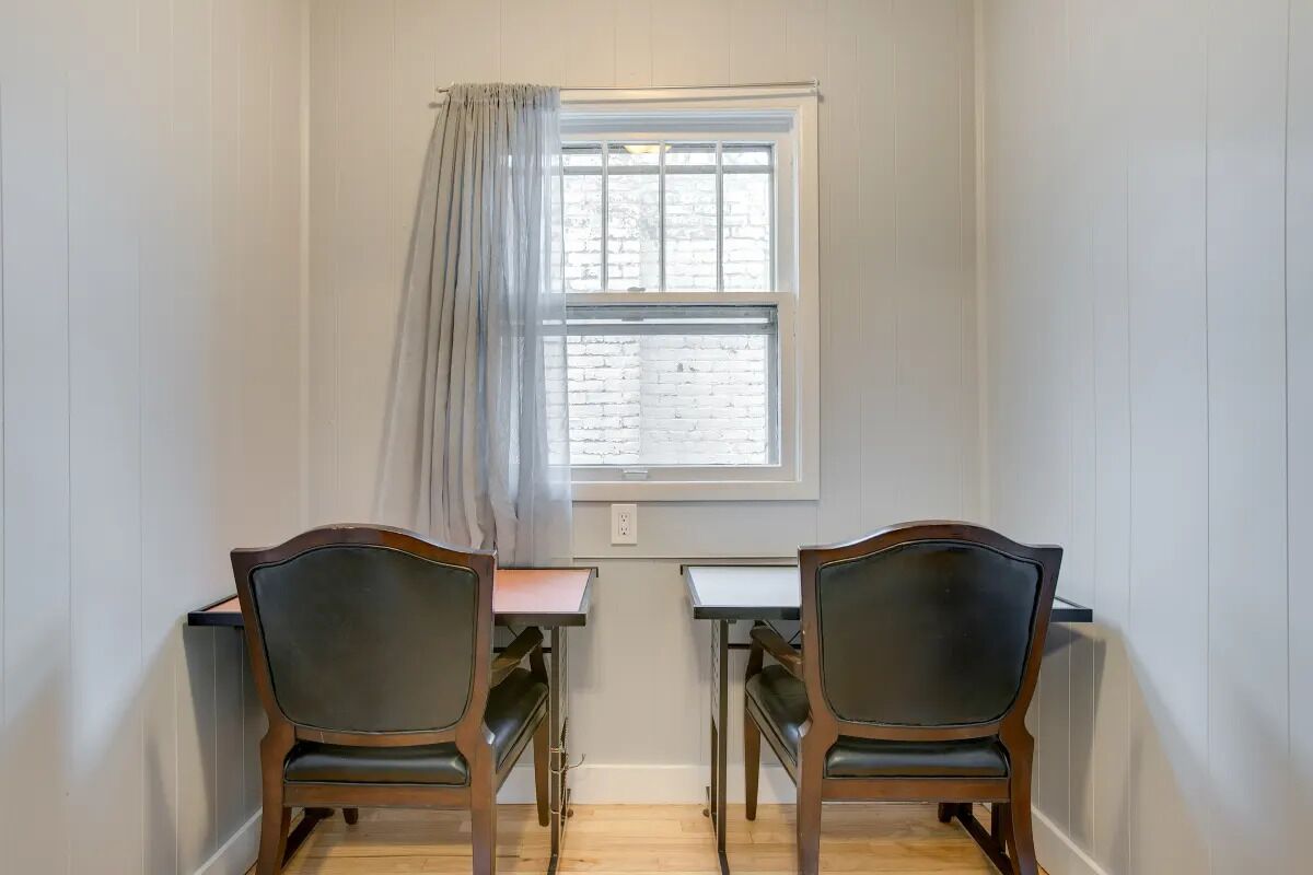 A room with two wooden chairs and a table, a window with a curtain, and a brick wall.