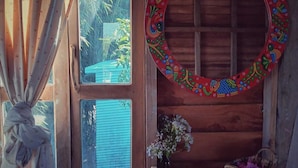 Interior de una cabaña de madera con ventana, un espejo decorativo y una pequeña estantería con un jarrón y una cesta.