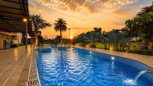 A swimming pool with a tiled edge, surrounded by palm trees and a sunset sky. - blue heaven resort jaipur  (Jamwa Ramgarh)