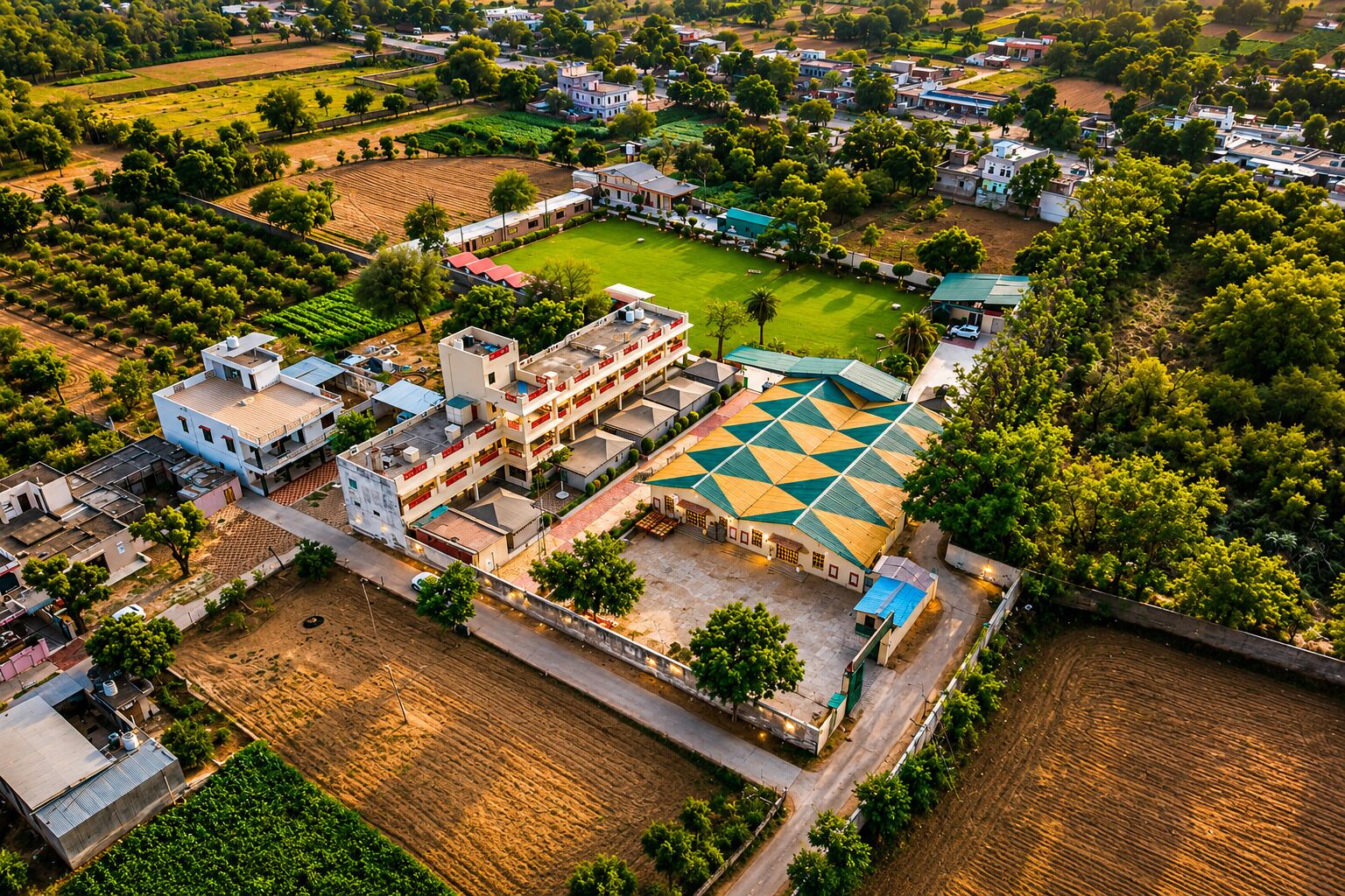 An aerial view of a residential area with a large building featuring a geometric roof pattern, surrounded by greenery and open fields.