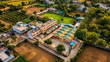 An aerial view of a residential area with a large building featuring a geometric roof pattern, surrounded by greenery and open fields.