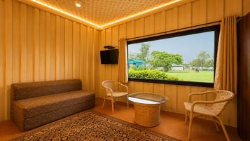 Una habitación con sofá, dos sillones de mimbre, una mesita redonda, televisor montado en la pared y una ventana con vistas a zonas verdes.