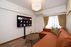 A living room with a flat-screen TV mounted on the wall, a brown sofa, a small round table with a vase, and a window with curtains.