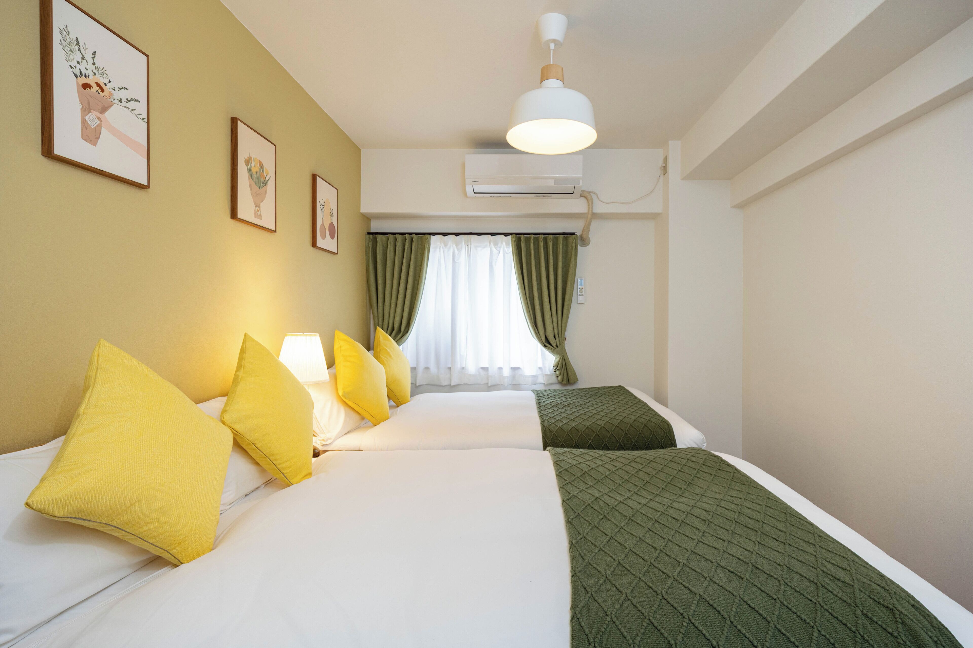 A hotel room with a bed, pillows, a lamp, an air conditioning unit, and a window with curtains.