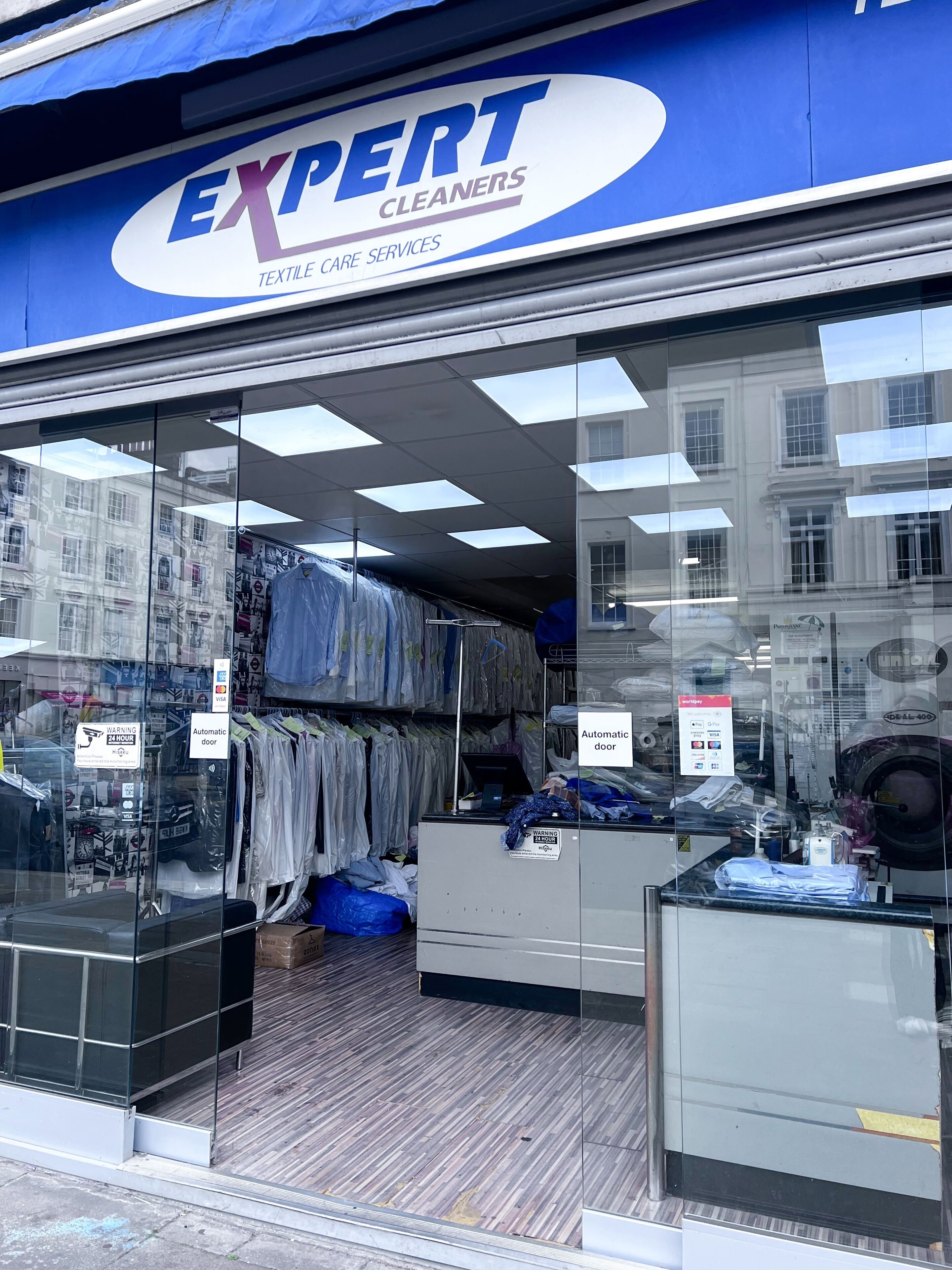 A storefront with a blue awning, glass doors, and a sign reading 'EXPERT CLEANERS TEXTILE CARE SERVICES.' Inside, there are shelves with folded clothes and a counter with cleaning supplies.