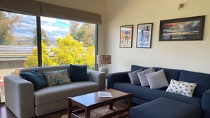 Ein Wohnzimmer mit zwei Sofas, einem Couchtisch, einem Teppich und großen Fenstern mit Blick auf Bäume und Hügel.