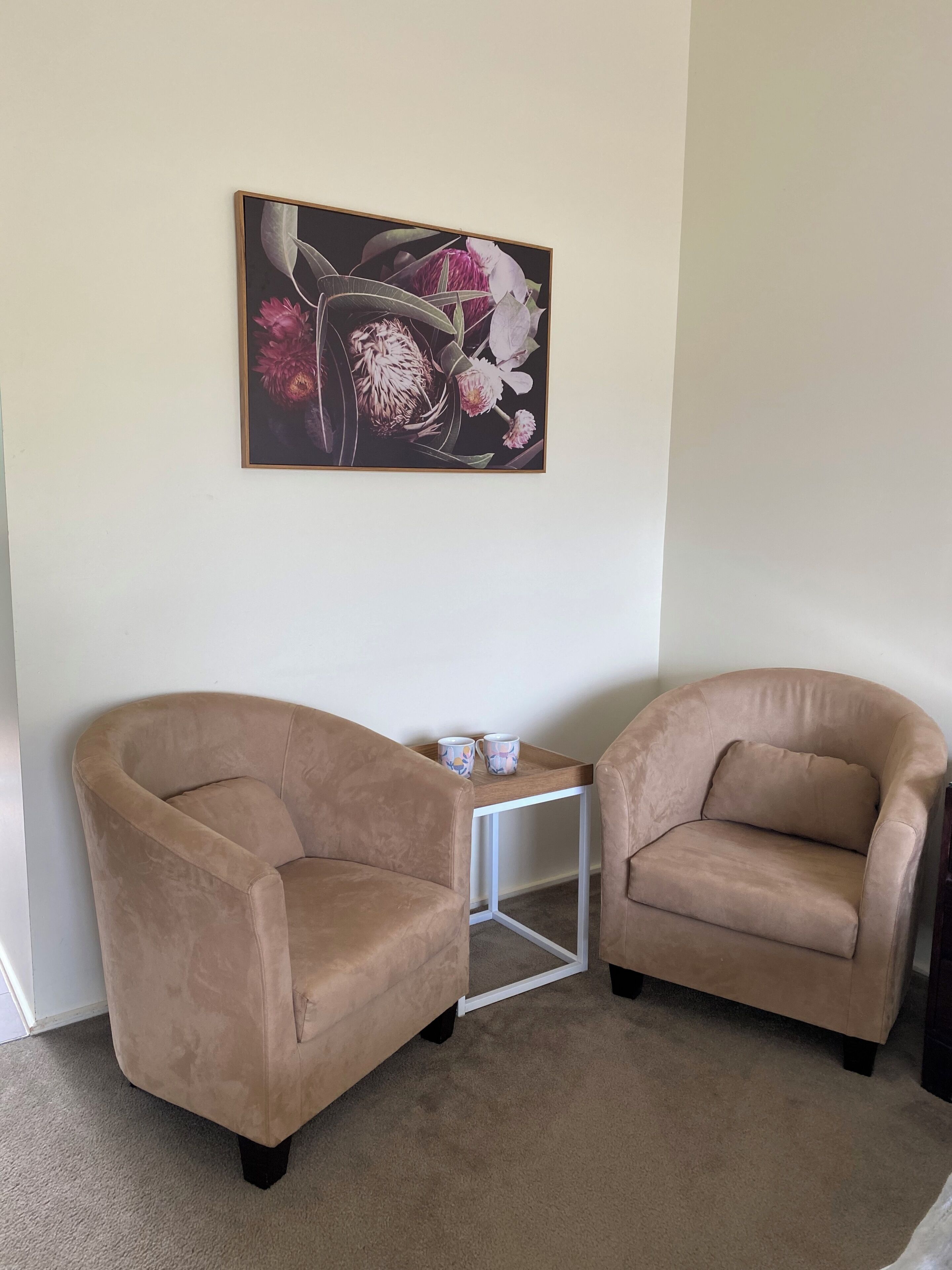 Two beige armchairs, a small white table with two mugs, and a floral painting on the wall.