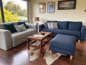 Un salon avec un canapé gris, un canapé bleu, une table basse en bois et un pouf bleu. Deux tableaux encadrés sont accrochés au mur, ainsi qu’une lampe et une grande fenêtre donnant sur des arbres.