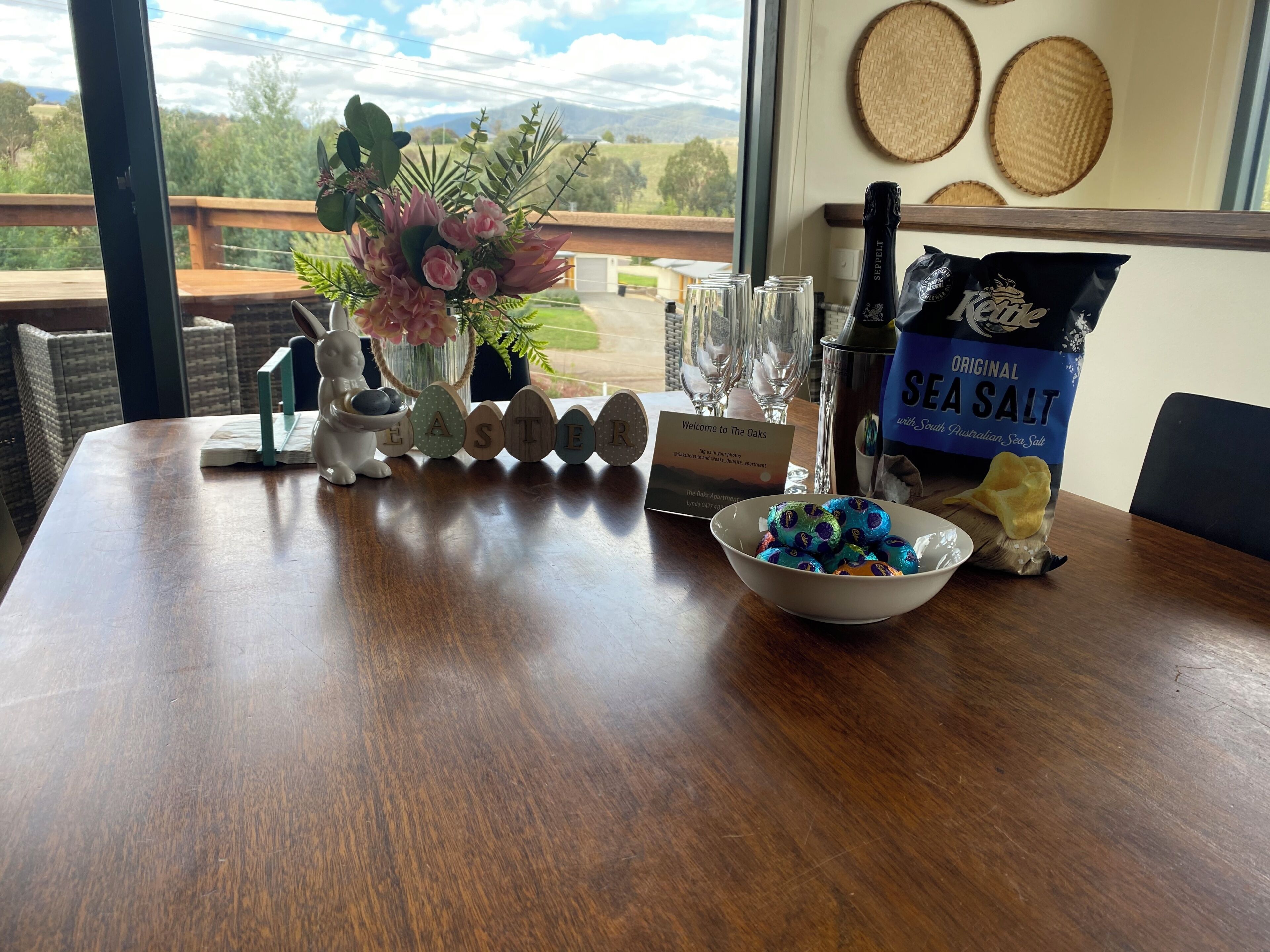 Une table en bois ornée d’un vase de fleurs, d’une bouteille de champagne, d’un bol de chocolats et d’une pancarte accueillant les clients de l’établissement The Oaks.