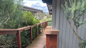 Eine Holzterrasse mit Geländer, umgeben von viel Grün und einem Gebäude im Hintergrund.