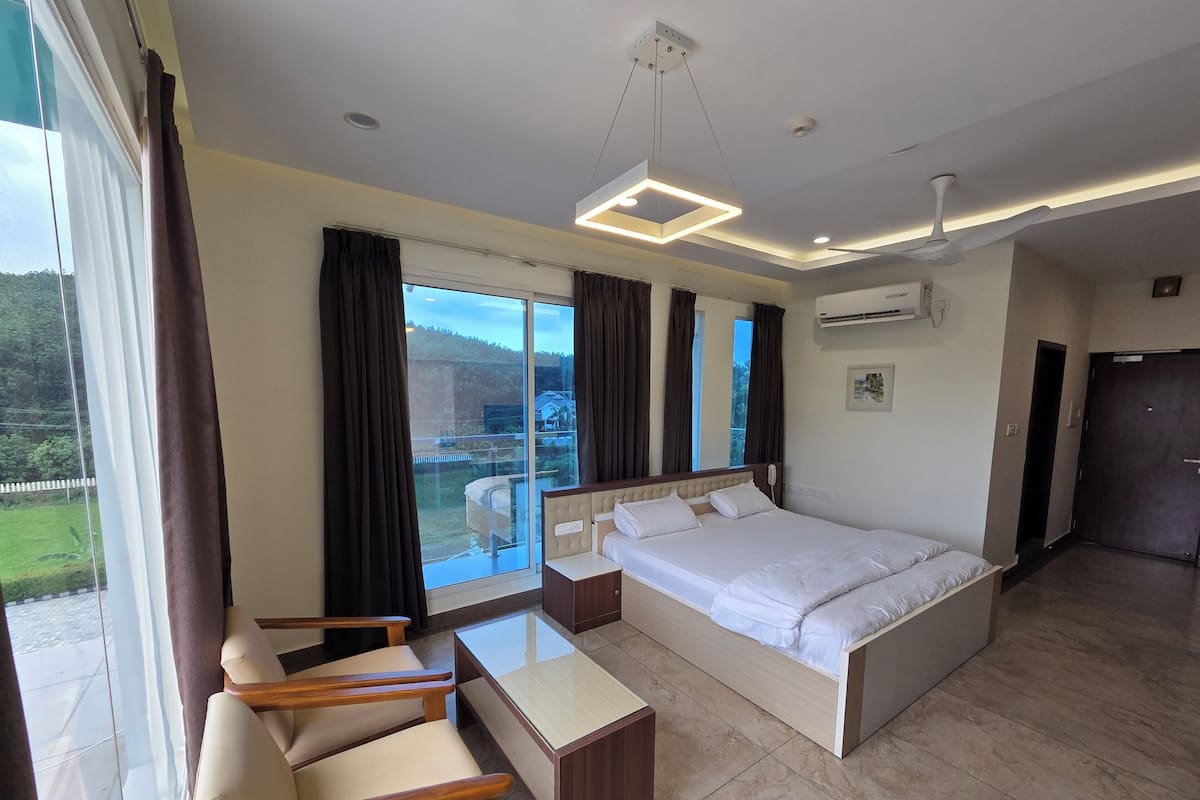 A modern hotel room with a large bed, a desk with chairs, a balcony with curtains, and a view of the outdoors.