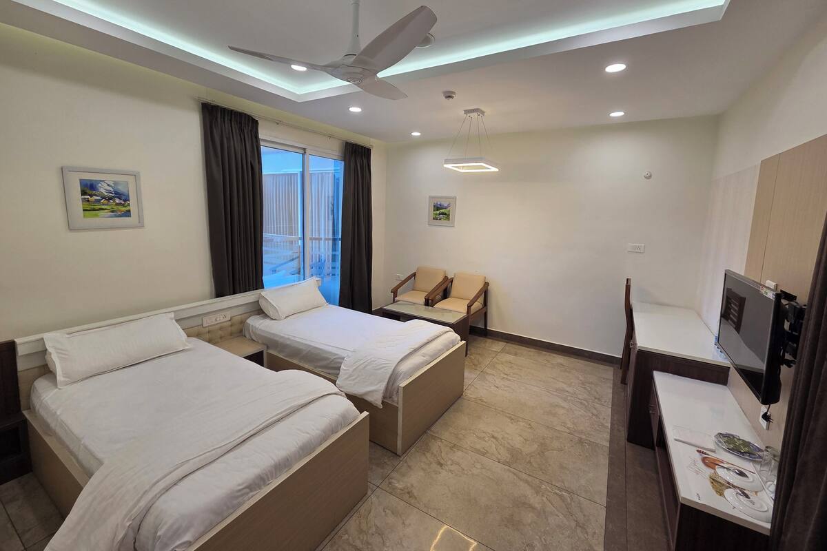 A hotel room with two beds, a desk, a chair, a ceiling fan, recessed lighting, and a window with curtains.