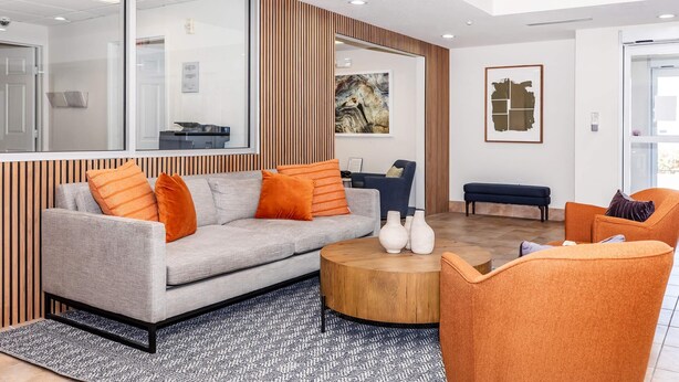 En moderne stue med en grå sofa, orange puder, et rundt træbord og en blå bænk.