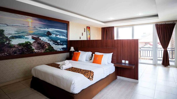 A hotel room with a large bed, a painting of a beach scene, a wooden headboard, and a view of the outside. - Nusa Dua Moon Hotel (Nusa Dua)
