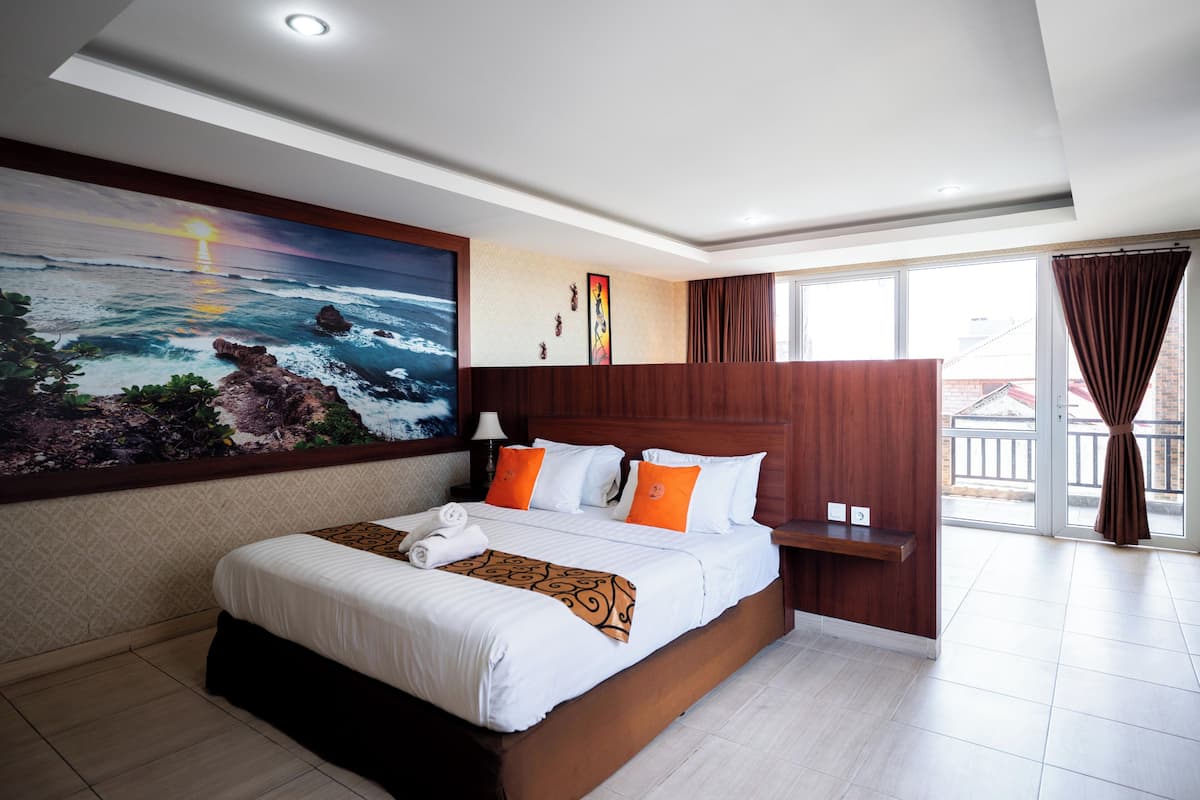 Ein Hotelzimmer mit einem großen Bett, einem Gemälde, das einen Sonnenuntergang über dem Meer zeigt, einem hölzernen Kopfteil und einem Balkon mit Vorhängen.