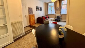 Una sala de estar moderna con una mesa de madera oscura, un sofá y una silla. Se observa un radiador, un aparador de madera y una ventana con cortinas.