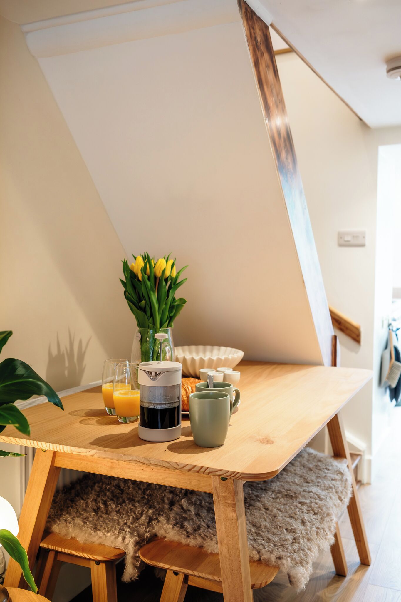 A wooden dining table with a vase of yellow flowers, a glass of orange juice, and a mug.