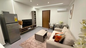 A modern living room with a flat-screen TV, a refrigerator, a sofa, a coffee table, and a dining area with a table and chairs.