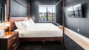 A modern bedroom with a four-poster bed, a nightstand, a bedside table with a lamp, and a large window offering a city view.