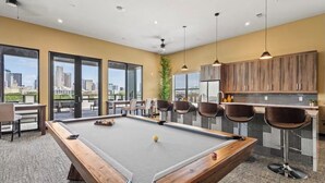 A pool table with chairs, a bar area, and large windows with a city view.