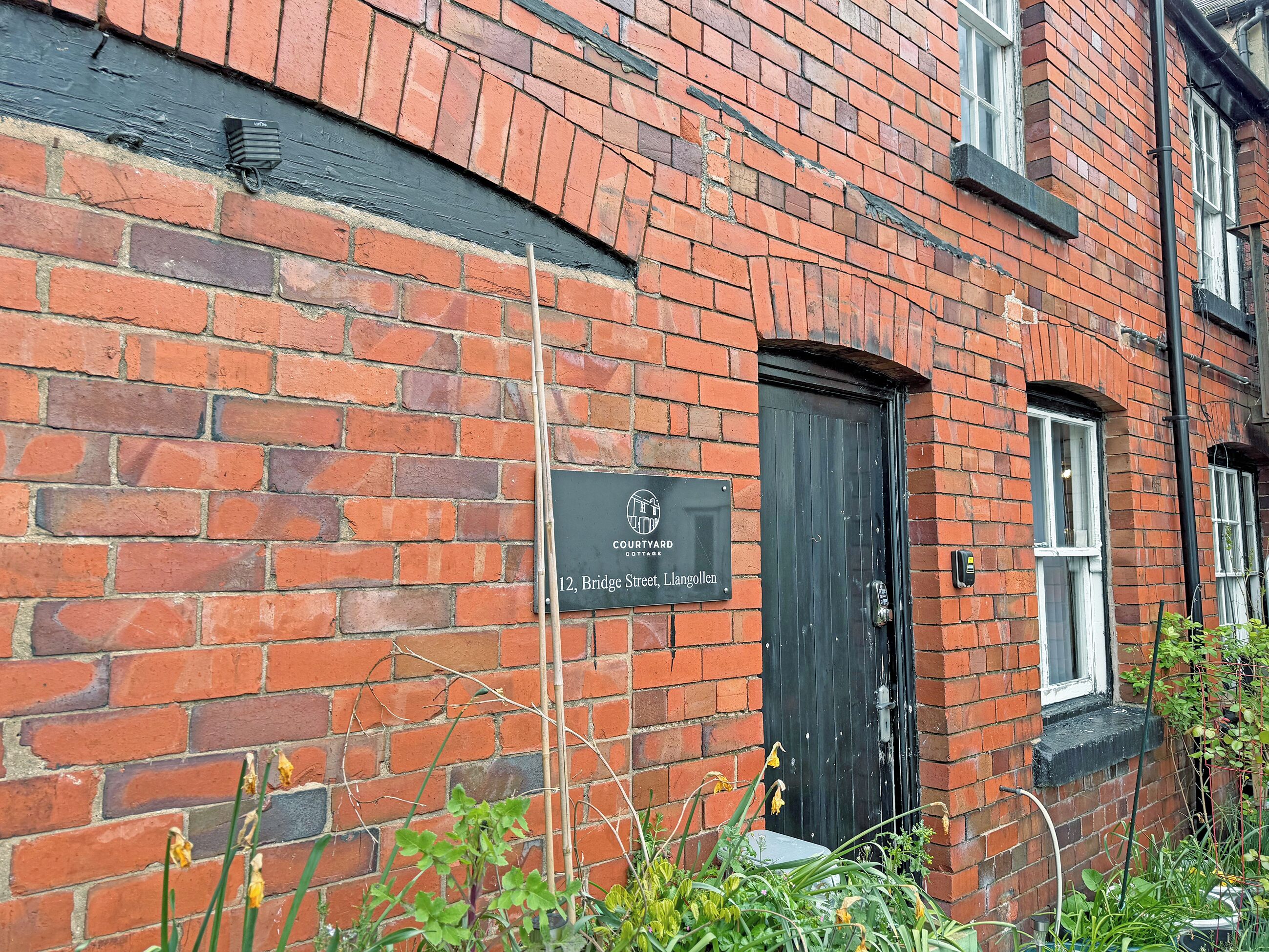 A brick building with a black door and a sign indicating 'Court Yard Cottage, 12, Bridge Street, Langollen.'